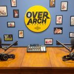 The OverArch Podcast studio in Vancouver, with two microphones and headphones set up on a wooden table beneath the OverArch wall logo.
