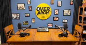 The OverArch Podcast studio in Vancouver, with two microphones and headphones set up on a wooden table beneath the OverArch wall logo.
