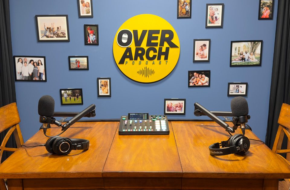 The OverArch Podcast studio in Vancouver, with two microphones and headphones set up on a wooden table beneath the OverArch wall logo.