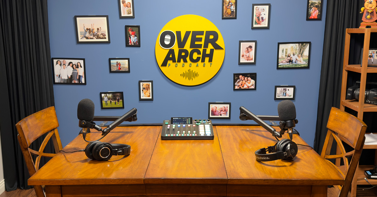 The OverArch Podcast studio in Vancouver, with two microphones and headphones set up on a wooden table beneath the OverArch wall logo.